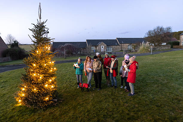 A wide, high-angle shot of a group of people standing together outside while singing Christmas carols. They are all standing next to a large Christmas tree, which is lit up with warm festive lighting. They are all wearing warm winter clothing with woolly hats. They are holding a booklet with Christmas carol lyrics. Everyone is smiling as they have come together as a community to celebrate Christmas.Videos are available similar to this scenario.