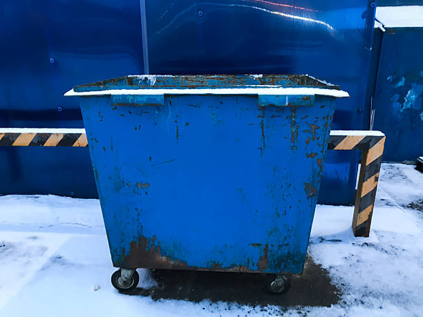 A blue dumpster with a yellow stripe on the side. It is parked on a snowy ground