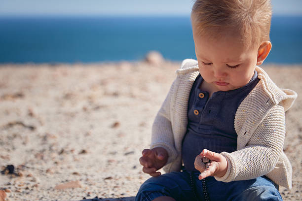 A lonely baby sits in the rocky sand by the sea. A sunny, relaxed moment by the ocean, full of charm and coastal vibes.