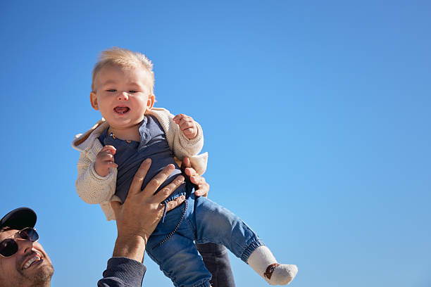 A joyful father-son moment as dad lifts his laughing baby into the air against a bright blue sky. A carefree, heartwarming scene full of love, trust, and play.