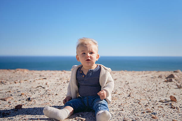 A lonely baby sits in the rocky sand by the sea. A sunny, relaxed moment by the ocean, full of charm and coastal vibes.