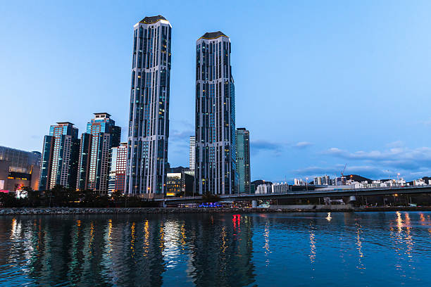 Busan urban skyline showcasing tall modern skyscrapers illuminated at dusk, reflecting on calm waterfront waters, backed by a serene evening sky. South Korea