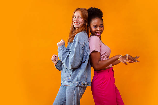 Two young multicultural girls, one with red hair and one with curly hair, pose joyfully in a vibrant studio with an orange backdrop; Their casual outfits reflect a fun and trendy Gen Z vibe