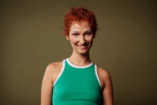 Portrait of a smiling girl with short red hair, wearing a green tank top, stands against a neutral olive-green background and looking at the camera