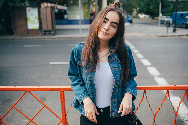 Woman in a blue jacket and white shirt is standing on a sidewalk next to a red fence