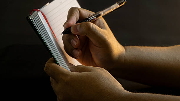 istock Side view of male hand holding upright notebook and pen under warm light, on black table with black background, cozy yet dramatic note-taking scene 2228640397
