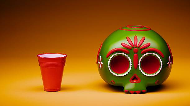 A glossy red plastic cup positioned next to a green ornamental sugar skull decorated with red and white floral motifs, set against a seamless orange background in a studio composition.
