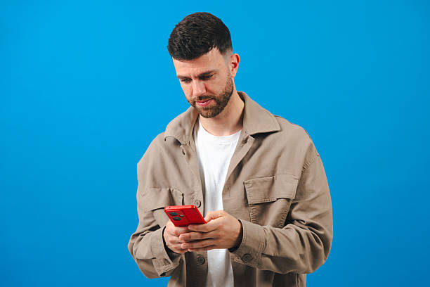istock Bearded young man texting on red smartphone, wearing jacket on a blue background 2228645259