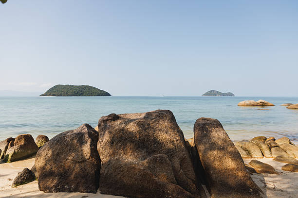 Beautiful tropical beach scene featuring large rocks on the shore and a calm sea with distant islands under a clear sky and sunlight, providing a peaceful and scenic view.