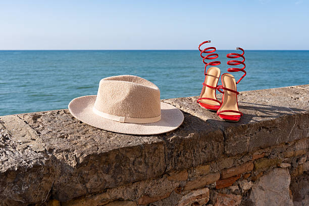 Elegant red high heels and a stylish straw hat placed on a stone wall with a breathtaking view of the sea, evoking a sense of summer vacation and relaxation