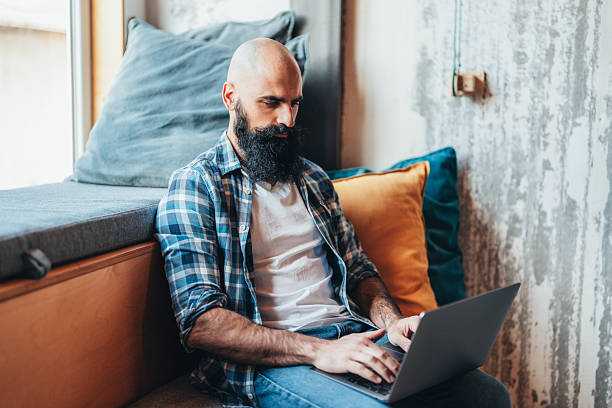 Serious man sitting and working on a laptop. He is wearing a white t-shirt and a blue plaid shirt, with a neatly groomed beard and a bald head. The man looks relaxed and content, while his hands are on the keyboard, mid-typing. In the background, there’s a softly lit, cozy space, perhaps at home or in a casual workspace.