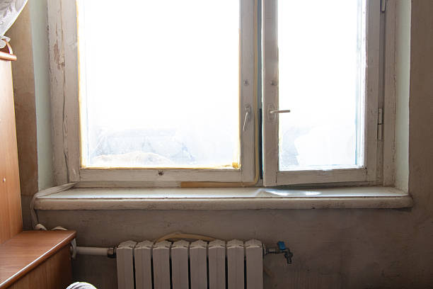 Old wooden window with foam insulation and peeling paint, under the window there is a heating battery on the wall in an apartment without renovation