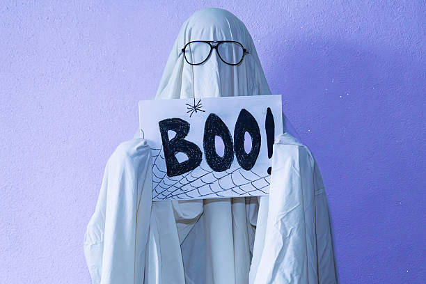A whimsical Halloween-themed image featuring a ghost costume with a white sheet and glasses holding a handwritten BOO! sign with cobweb doodles. Shot against a purple textured background, it delivers playful spooky vibes.