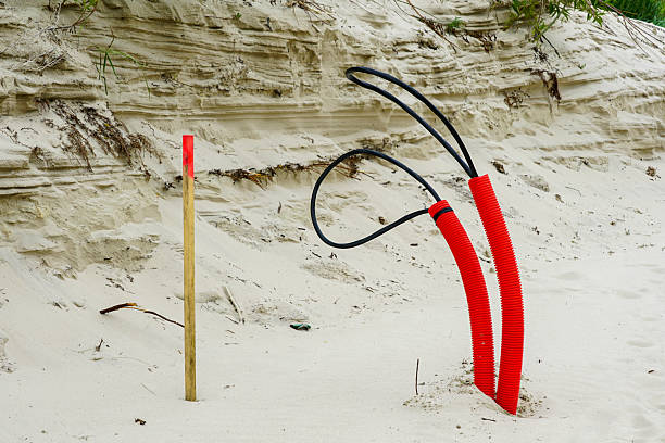 Bright red electrical conduit pipes and a wooden marker stake emerge from sand at a infrastructure construction site by a beach dune