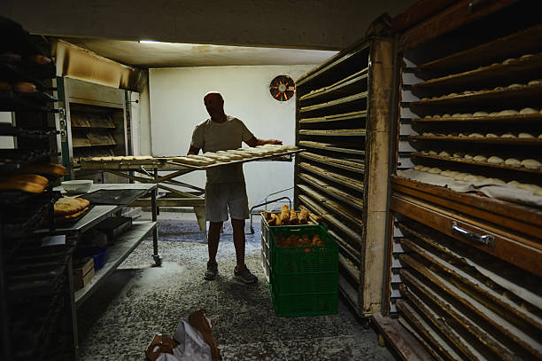 Artisan baker organizing dough and baked breads in a rustic bakery setting with traditional ovens and shelves filled with bread trays.