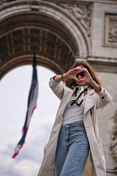 Smiling woman in Paris forming heart shape with hands in front of Arc de Triomphe and waving French flag
