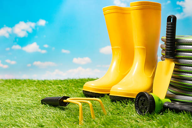 Gardening and spring work concept made with bright yellow rubber boots and small garden tools next to coiled watering hose on a vibrant green lawn under a blue sky.