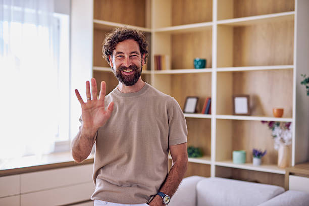 istock Smiling man waving hand looking at camera in living room 2228688877
