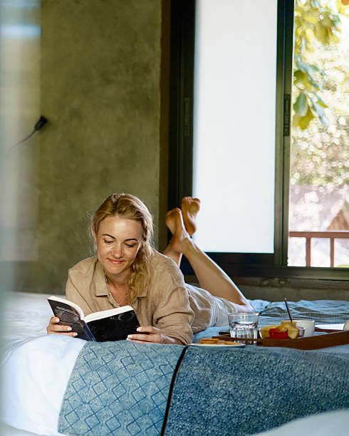 istock Smiling woman is reading a book while lying on a bed next to breakfast. 2228690614