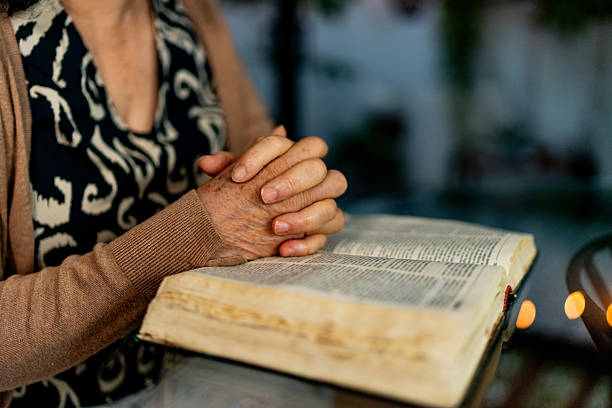 Pray and bible concept. Hand in hand together on bible by senior woman (worship christian), thinking and closed her eyes at church. Person side view, female praying and hope for peace-vertical image.