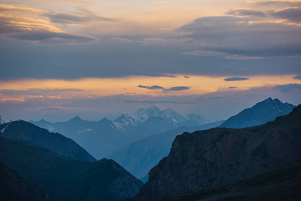 Deep gorge between layered silhouettes of sharp rocky ridge spurs under huge snowy mountain peak in golden cloudy sky. Sheer crags of mount valley under giant ice top under gold sunset color clouds.