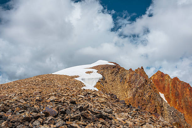 istock Snow-capped mountain ridge with cornice and red sharp rocks on top in sunlight. Snow cap and shiny pointy peak of gold color. Snow dome and colorful rocky peaked top. Sunny cloudy high mountains view. 2228702154