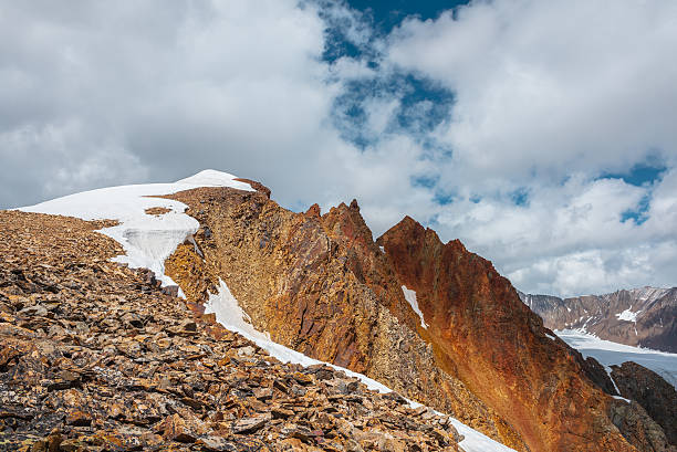 istock Snow-capped mountain ridge with cornice and red sharp rocks on top in sunlight. Snow cap and shiny pointy peak of gold color. Snow dome and colorful rocky peaked top. Sunny cloudy high mountains view. 2228702156