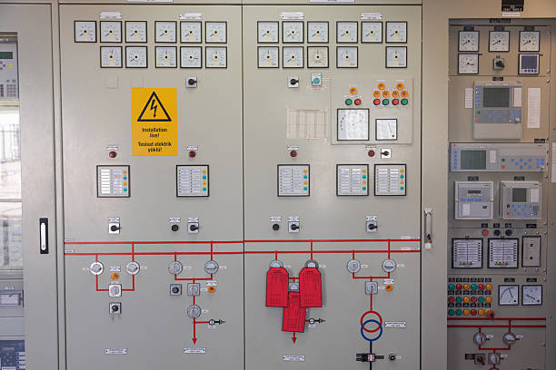 A detailed view of a high voltage electrical control panel inside a modern power plant control room. The panel features multiple analog meters, warning lights, digital displays, push buttons, rotary switches, and a prominent yellow warning sign that reads \