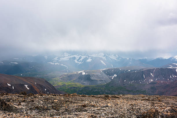 istock Dramatic top view from sunlit stony pass to vivid green alpine valley in sunlight against large mountain range with snow-capped peaks in rainy low clouds. Misty snowy mountain tops in rain cloudy sky. 2228714868