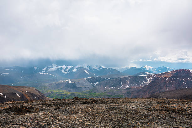 istock Dramatic top view from sunlit stony pass to vivid green alpine valley in sunlight against large mountain range with snow-capped peaks in rainy low clouds. Misty snowy mountain tops in rain cloudy sky. 2228714872