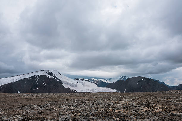 istock Dramatic misty top view from stone pass to big glacier tongue among sharp rocks and large snow-capped pinnacle in rainy low clouds. Dark atmospheric mountain silhouettes in rain in gray cloudy sky. 2228714892