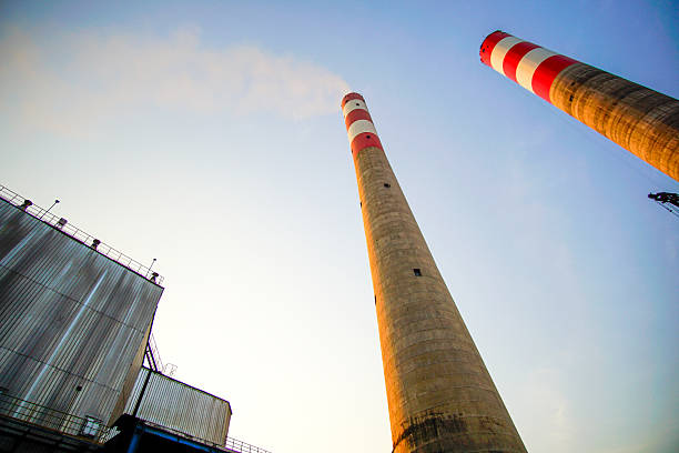 A low-angle shot captures the imposing smokestacks of an industrial plant against a clear sky, evoking a sense of scale and the impact of industry on the environment.