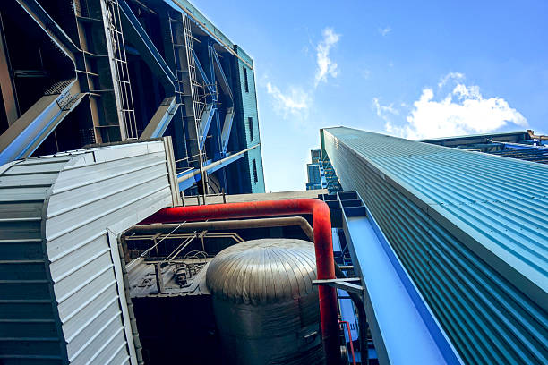 A low-angle shot captures the impressive scale of industrial architecture, with metal structures and piping set against a bright blue sky dotted with clouds