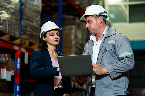 istock White adult female logistics staff using laptop beside storage racks during warehouse task operation, wearing safety helmet and navy blazer focused on screen, inside a logistics warehouse. 2228721250