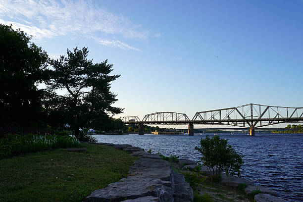 Partial view of a bridge spanning the Ottawa River, as seen from a grassy shoreline with trees in the foreground