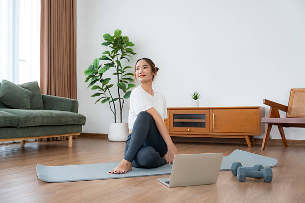 istock Young Asian woman stretching yoga workout on exercise mat while online training class with computer laptop in living room 2228725014