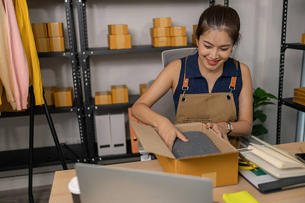 istock Smiling young female entrepreneur packing clothes into a parcel box, happily preparing merchandise orders, managing her successful online fashion store from her organized home workspace. 2228726114