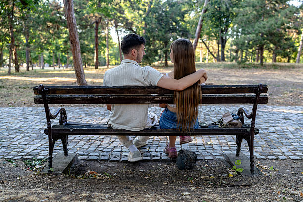 A couple shares a tender embrace while seated on a rustic park bench surrounded by greenery in a public park. Captured from behind, the image focuses on their closeness and connection, evoking a sense of intimacy, warmth, and serenity. Their relaxed posture and the tranquil surroundings highlight the bond they share.