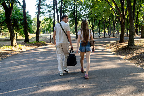 A serene scene featuring a young couple strolling through a sunlit park under tall trees. Carrying bags, they walk closely together, engaged in conversation. The warm sunlight and picturesque surroundings create an atmosphere of relaxation and togetherness. Their movement conveys companionship and the joy of being outdoors in pleasant weather.