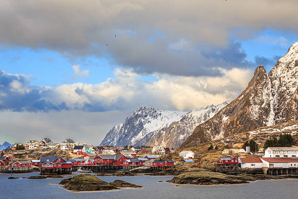 Picturesque fishing village on Lofoten Islands, Norway, with red cabins, snowy mountains, dramatic sky, and rugged coastline, traditional Nordic scenery.