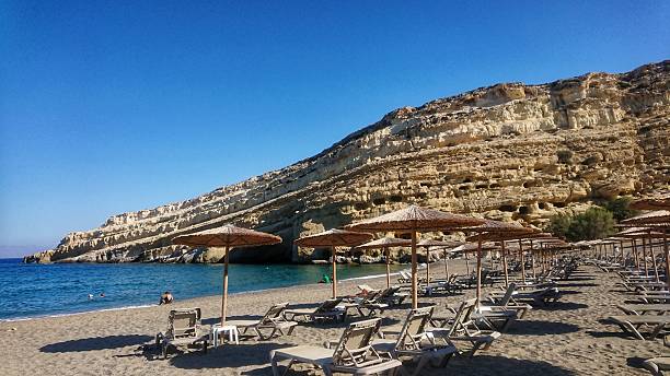 Scenic view of Matala Beach on Crete Island, Greece. Rows of sunbeds and straw umbrellas facing the turquoise sea and dramatic limestone rock formations.