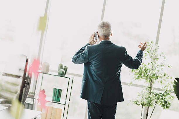 Confident senior businessman using a mobile phone in a modern office setting with large windows and lush greenery