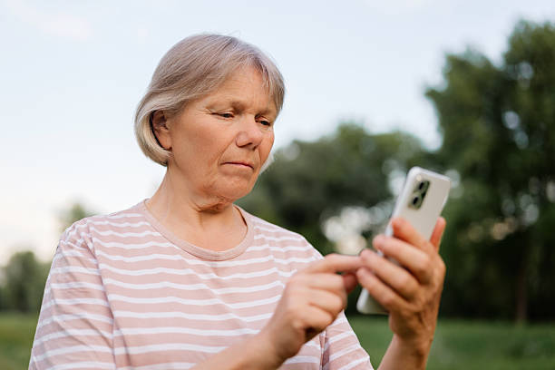 istock A middle-aged woman with light, short hair stands outdoors, intently focused on interacting with her smartphone, with a blurred green natural background. 2228744642