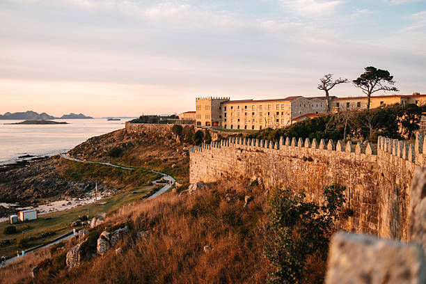 Scenic coastal landscape featuring the stone walls of Baiona’s fortress overlooking the sea and rocky shoreline in warm evening light.