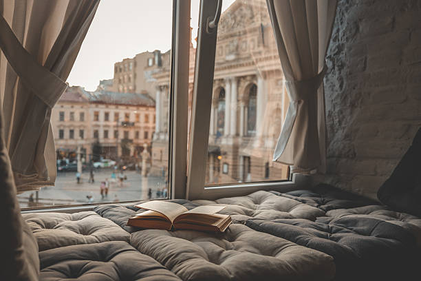 istock Cozy reading nook with an open book on a cushioned window seat overlooking a historic European city square and opera house at sunset through soft curtains 2228747011