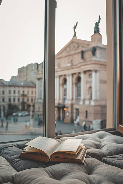 istock Cozy reading nook with an open book on a cushioned window seat overlooking a historic European city square and opera house at sunset through soft curtains 2228747012
