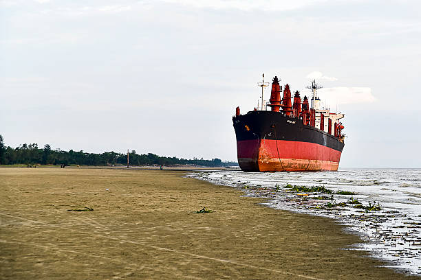 Chittagong, Bangladesh - 20 June, 2017: A large cargo ship rests beached along the sandy shoreline in Bangladesh, creating a striking maritime scene. The red and black hull of the vessel contrasts sharply with the muted tones of the sand and sky, while gentle waves lap against its side. This image reflects the shipbreaking and maritime heritage of Bangladesh’s coastal regions, known for being home to some of the world’s largest ship recycling yards. The vast beach and distant greenery add a sense of scale and atmosphere to the composition, making it a captivating subject for photography, industry documentation, and travel imagery.
