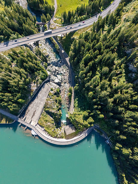 Aerial view of a curved dam holding turquoise water surrounded by dense green forest. Vehicles travel on a highway bridge above the dam. Scenic landscape with natural and man-made elements.