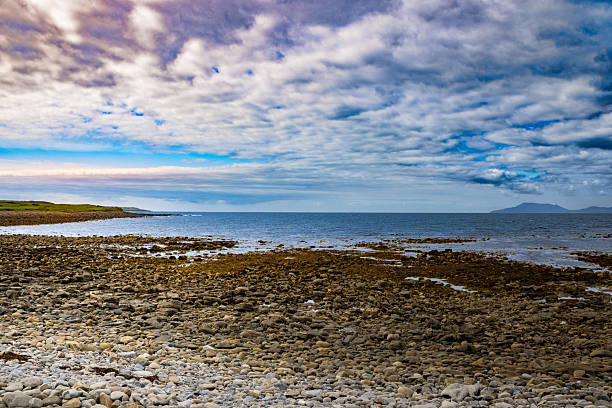 A wide view of the rocky shoreline at Tullaghan Beach, County Leitrim, with scattered seaweed and stones leading into the Atlantic. Distant mountains rise under a dramatic cloud-filled sky.