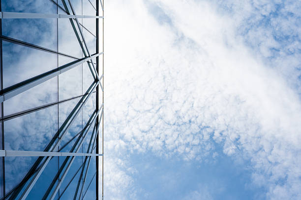 Contemporary glass building facade with geometric lines reflects a bright blue sky and scattered clouds. Suitable for illustrating architecture, urban design, or business concepts in presentations and marketing materials.
